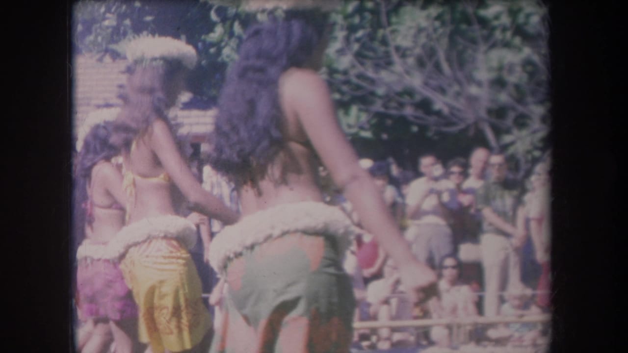 1969 Hawaii 8mm frame with hula dancers wearing leis and skirts, seen from behind and side-on before a shaded audience.