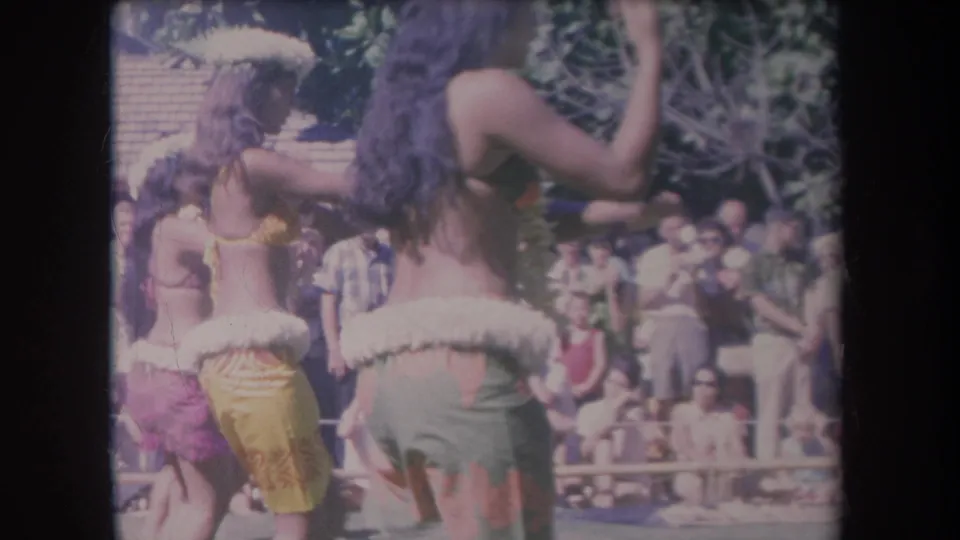1969 Hawaii 8mm frame showing dancers mid-routine, with raised arms, skirts, leis, and spectators in the background.