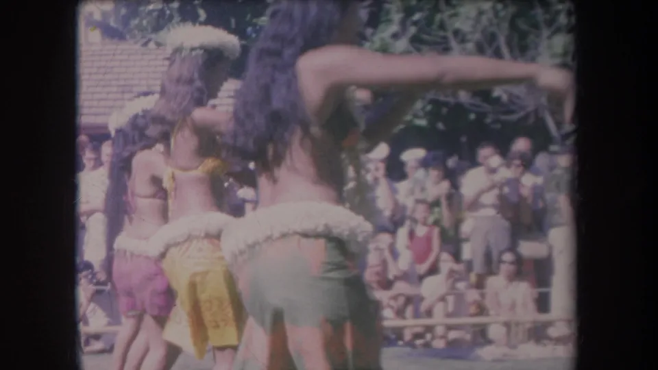 1969 Hawaii 8mm frame with a dancer's outstretched arm in the foreground, other dancers behind, and an outdoor audience watching.