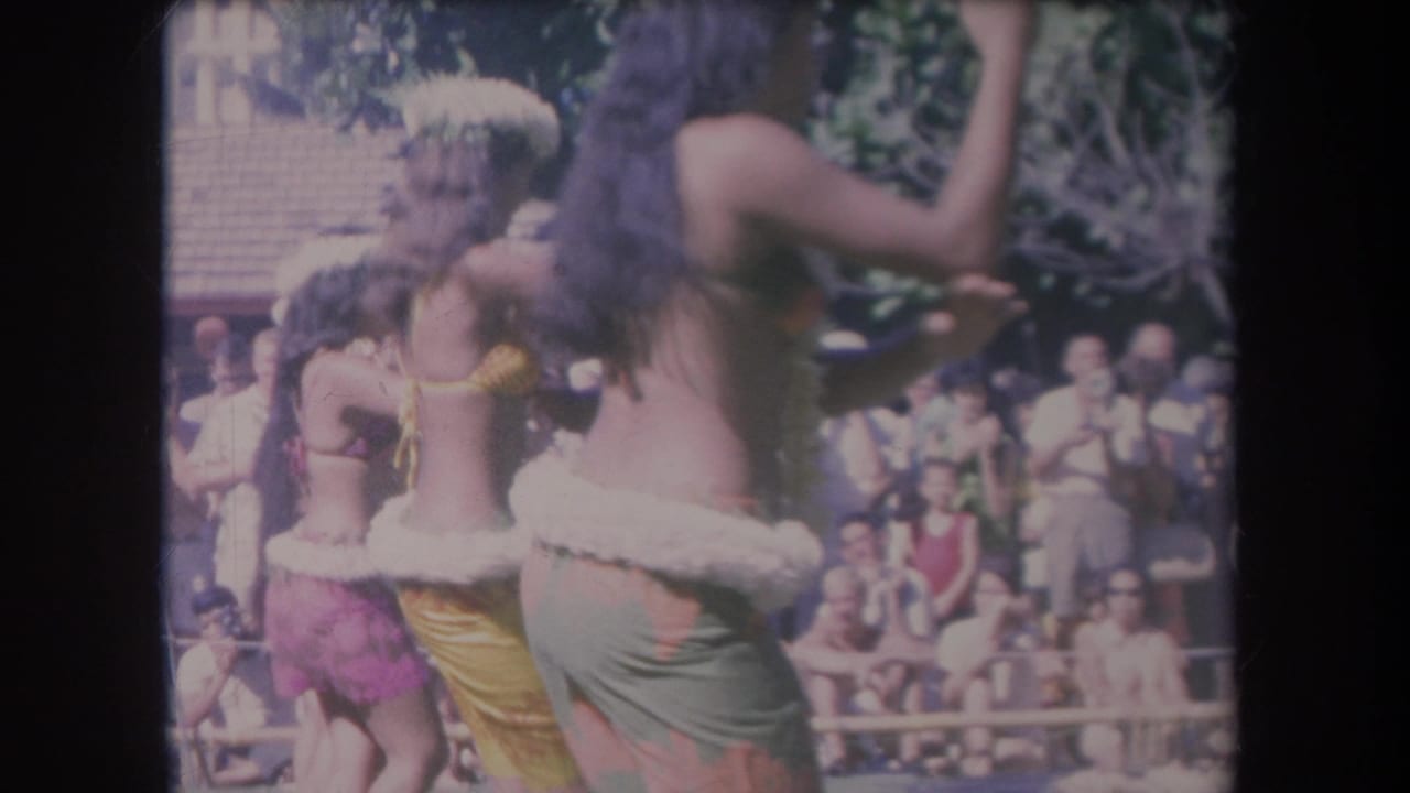 Hula dancers perform outdoors before a seated crowd in bright sunlight.