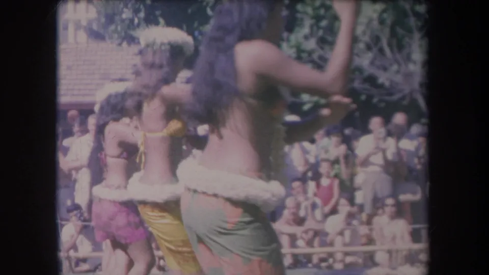 1969 Hawaii 8mm frame showing hula dancers in leis and skirts performing for tourists near trees and a wooden structure.