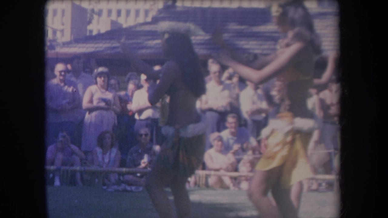 1969 Hawaii travel footage frame with hula performers mid-gesture in front of a tourist audience and rustic building.