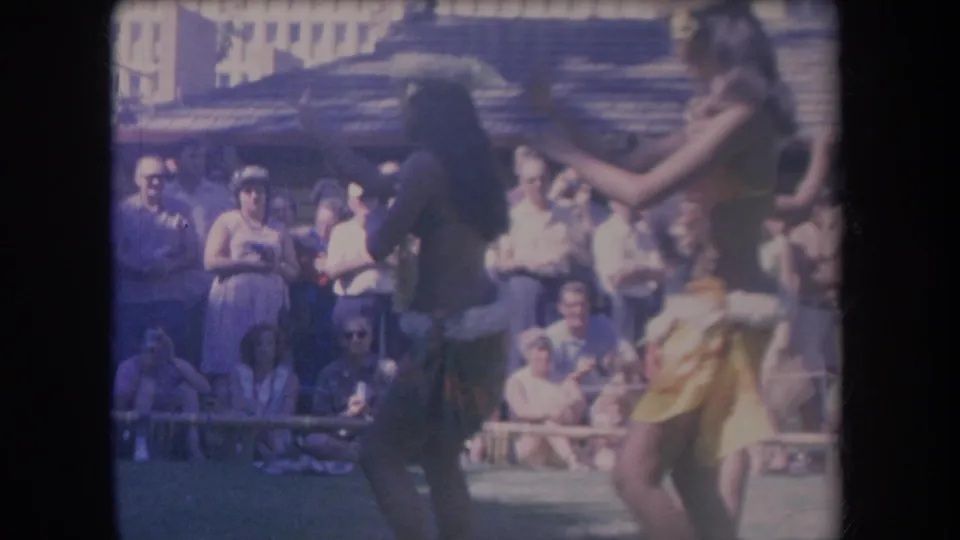 1969 Hawaii travel footage frame with hula performers mid-gesture in front of a tourist audience and rustic building.