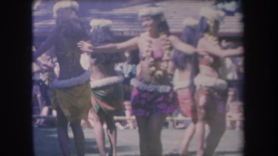 1969 Hawaii 8mm frame showing performers in leis and colorful skirts dancing near a crowd at an outdoor attraction.