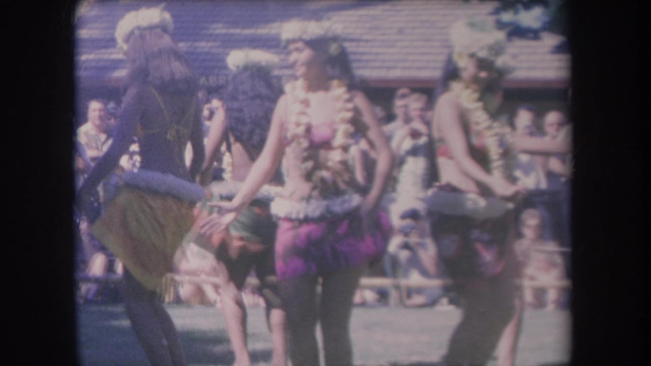 1969 Hawaii home-movie frame showing hula dancers in leis and skirts moving in front of spectators and a wooden-roofed structure.