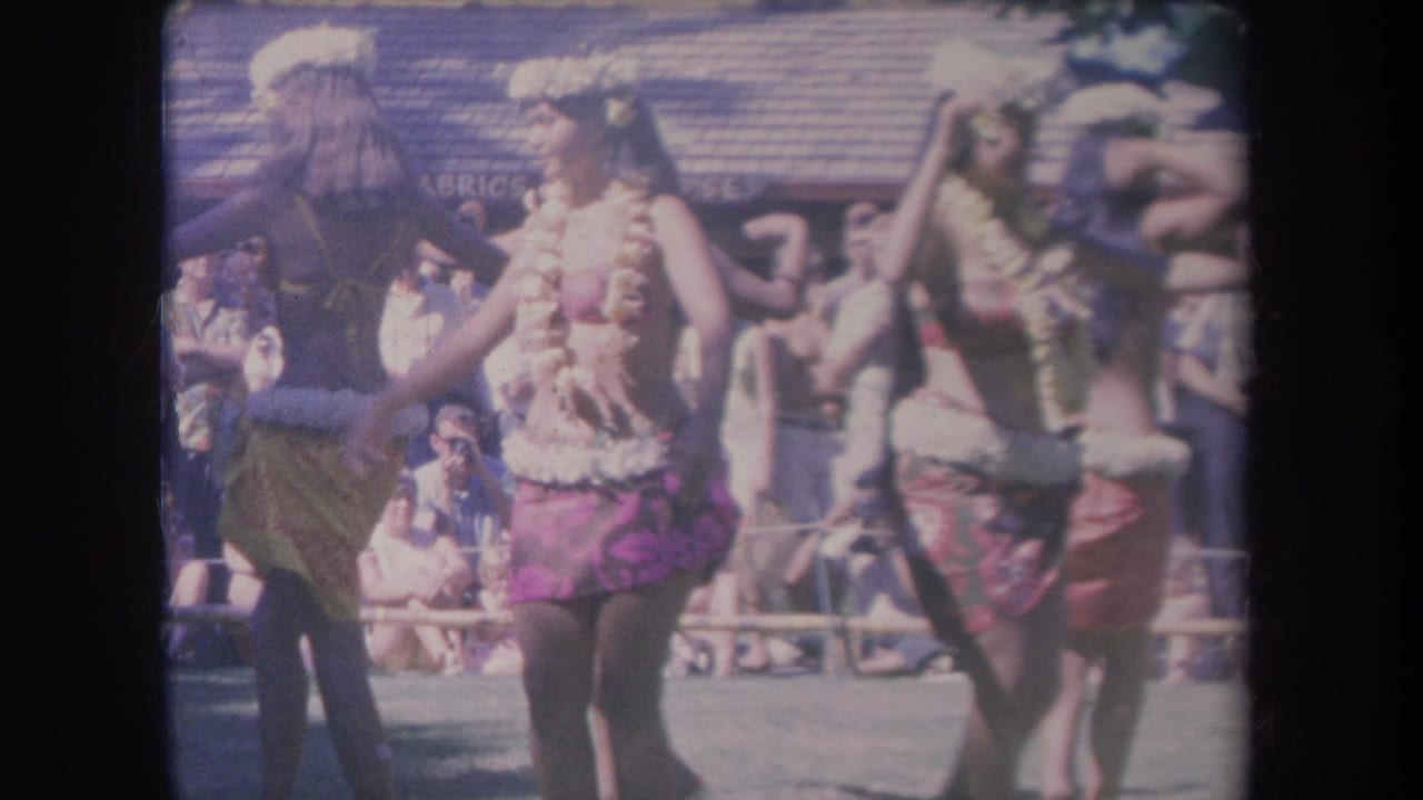 1969 Hawaii frame of several hula dancers in floral leis and skirts, with spectators and partial background signage visible behind them.