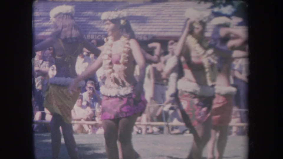 1969 Hawaii frame of several hula dancers in floral leis and skirts, with spectators and partial background signage visible behind them.
