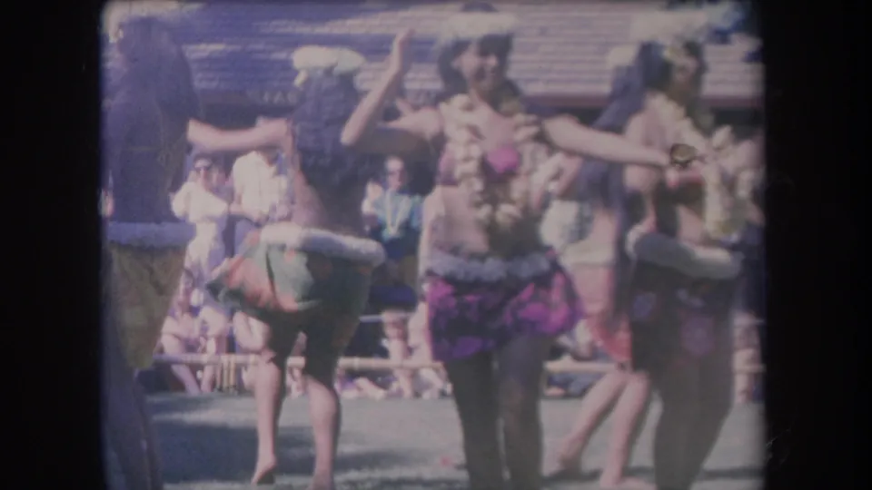 1969 Hawaii home-movie frame with a central dancer in lei and purple skirt, other performers nearby, and audience members seated behind.