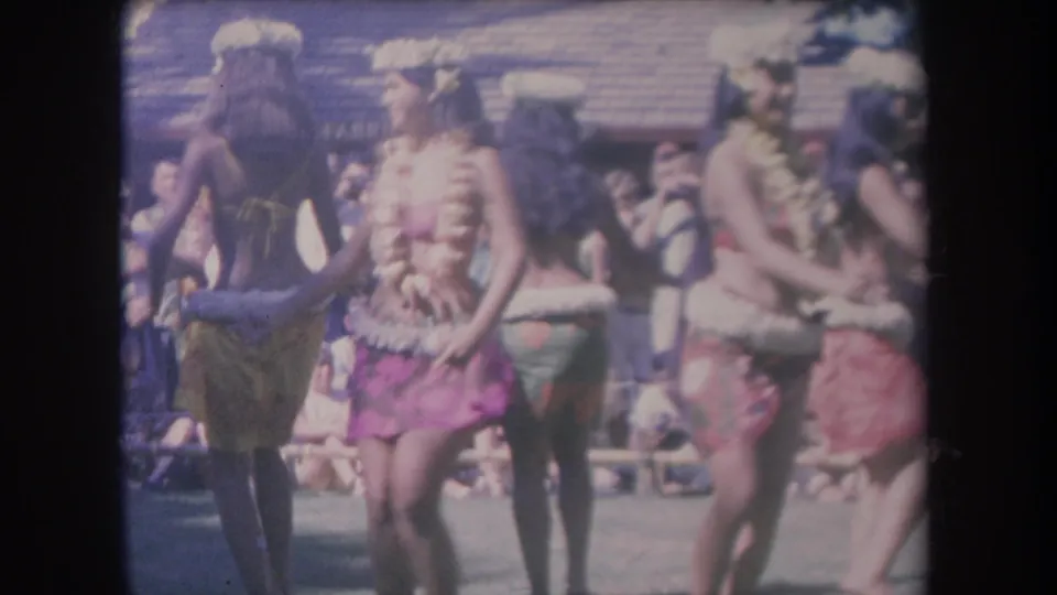 1969 Hawaii frame showing dancers in leis, skirts, and headpieces during an outdoor hula performance before a seated audience.
