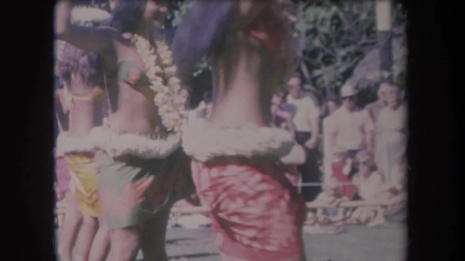 1969 Hawaii home-movie frame showing young women hula dancing in leis and skirts before an outdoor audience.