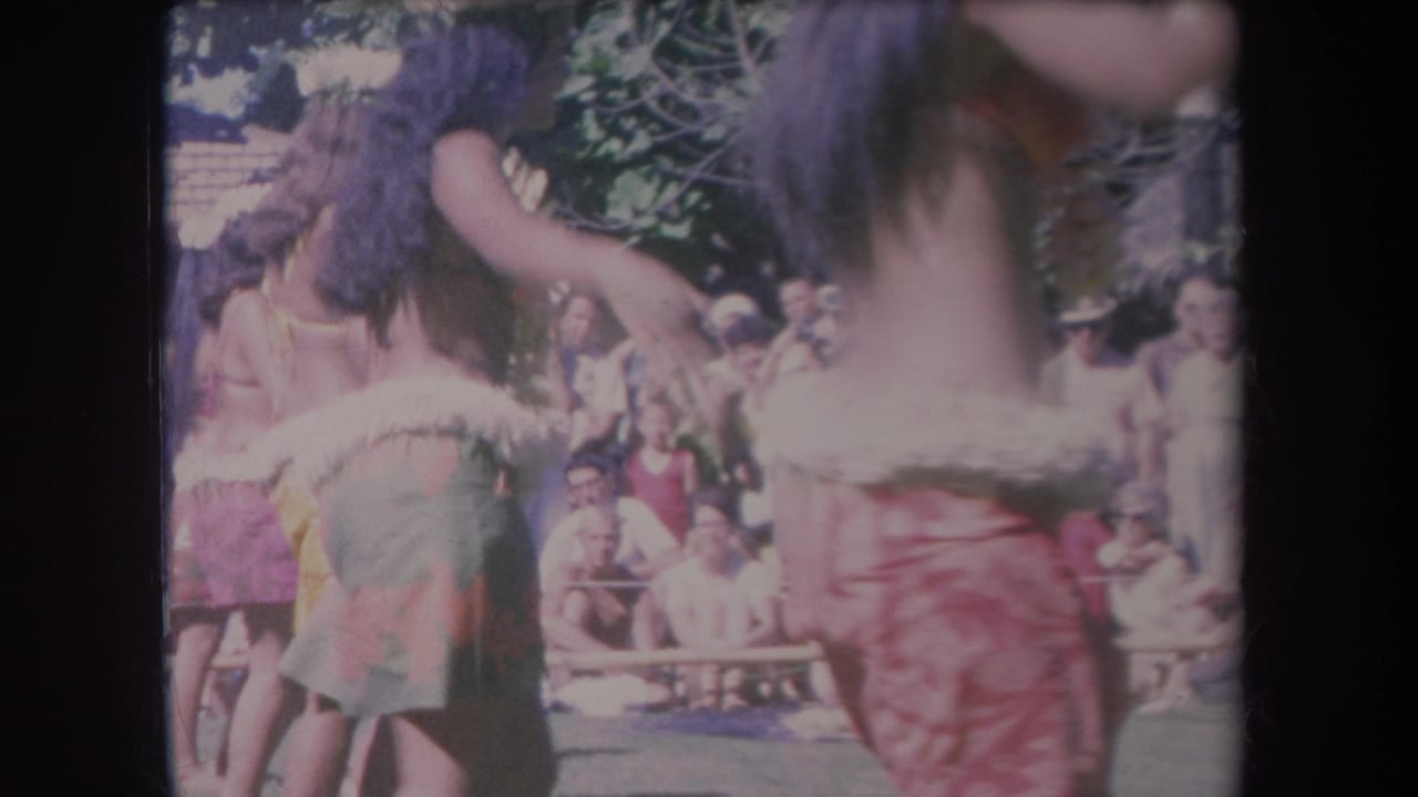1969 Hawaii 8mm frame of hula dancers in tropical attire moving past a seated outdoor audience.
