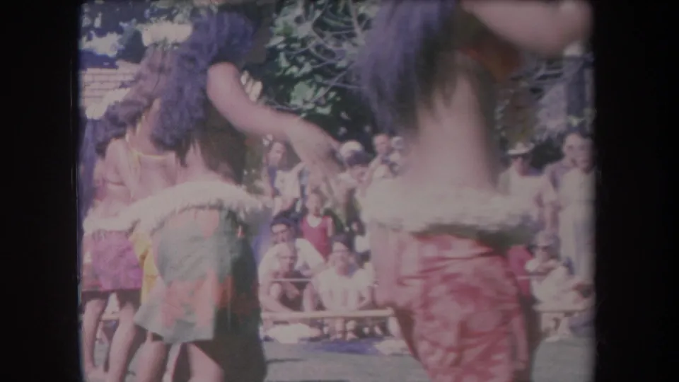 1969 Hawaii 8mm frame of hula dancers in tropical attire moving past a seated outdoor audience.