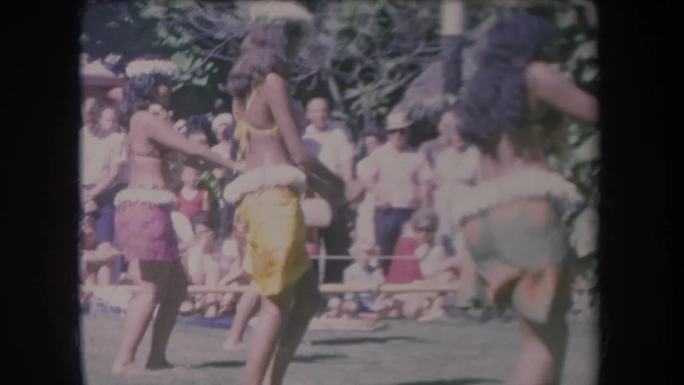 1969 Hawaii home-movie frame with barefoot hula dancers in yellow, purple, and green costumes before an audience.