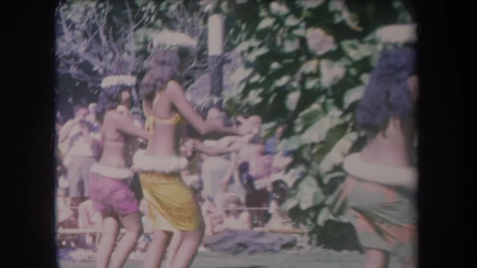 1969 Hawaii 8mm frame showing hula dancers from behind with a seated crowd and tropical greenery nearby.