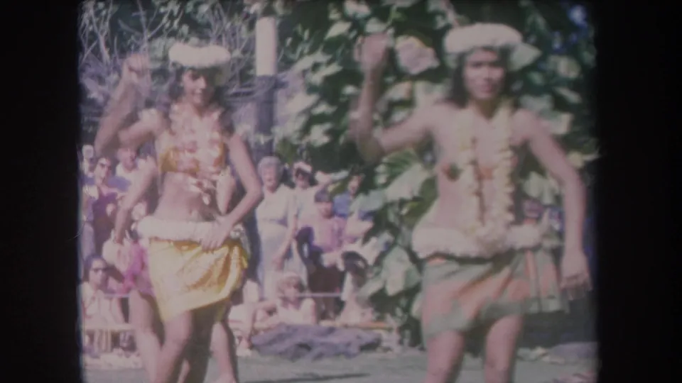 1969 Hawaii frame showing two women in traditional-style dance costumes performing outdoors before an audience.