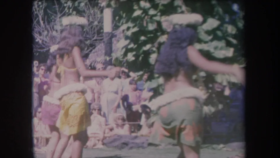 1969 Hawaii home-movie frame of dancers moving across an outdoor stage area in front of spectators.