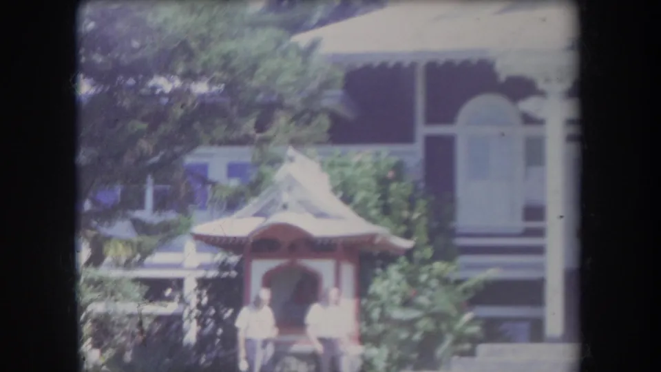 A red-and-white gazebo sits in a Hawaii residential garden as two distant people stand near house steps and surrounding foliage.