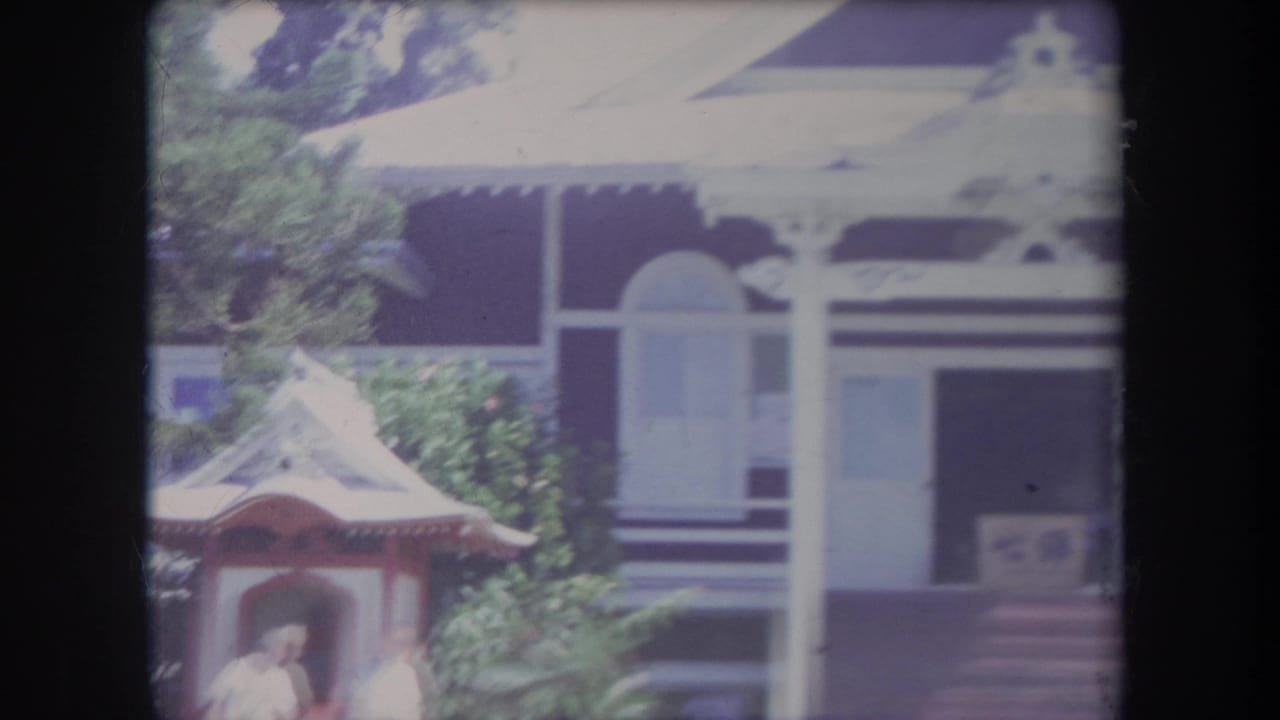 1969 Hawaii 8mm frame showing a gabled house with white trim, covered porch, garden feature, greenery, and a person in light clothing.