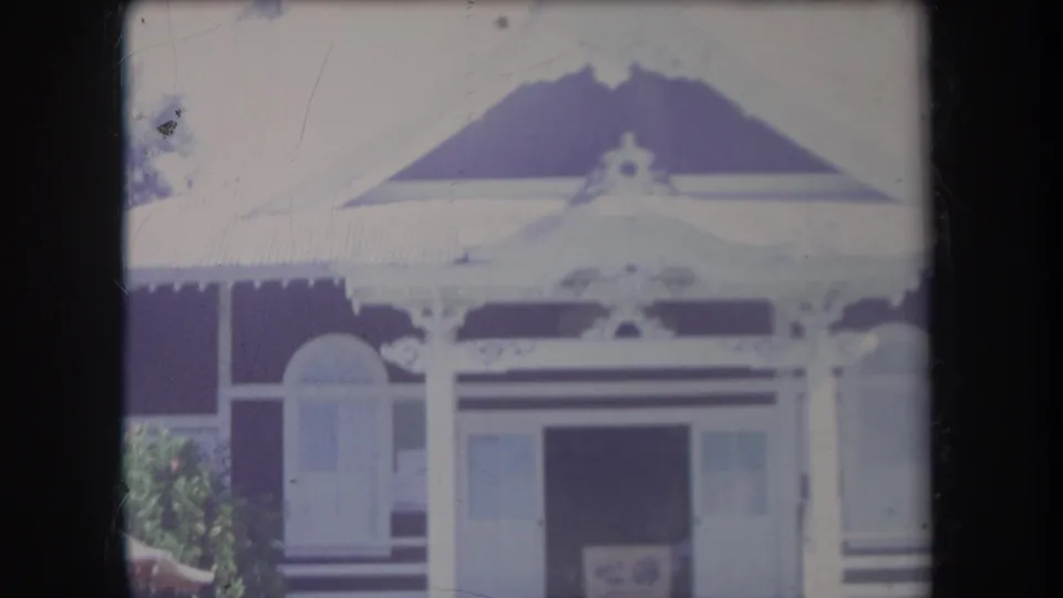 1969 Hawaii home-movie frame of a decorative house front with gabled roof, white trim, porch posts, and an open central doorway.
