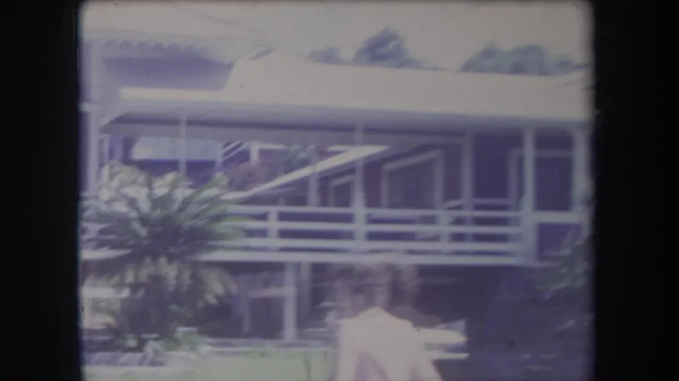 1969 Hawaii home-movie frame showing a covered side porch or veranda with railings, tropical plants, and a person in light clothing.