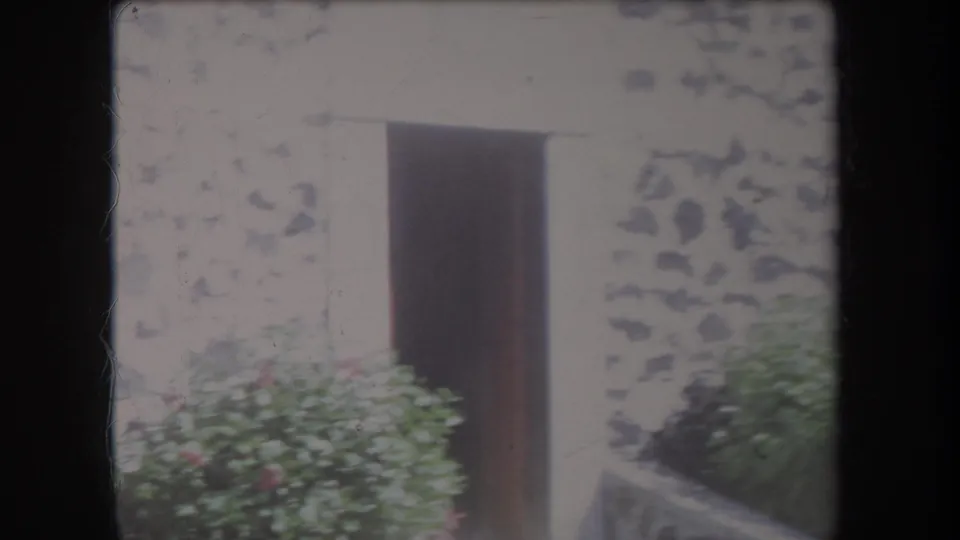 1969 Hawaii home-movie frame showing an open doorway, weathered cement facade, and shrubs near the entrance.