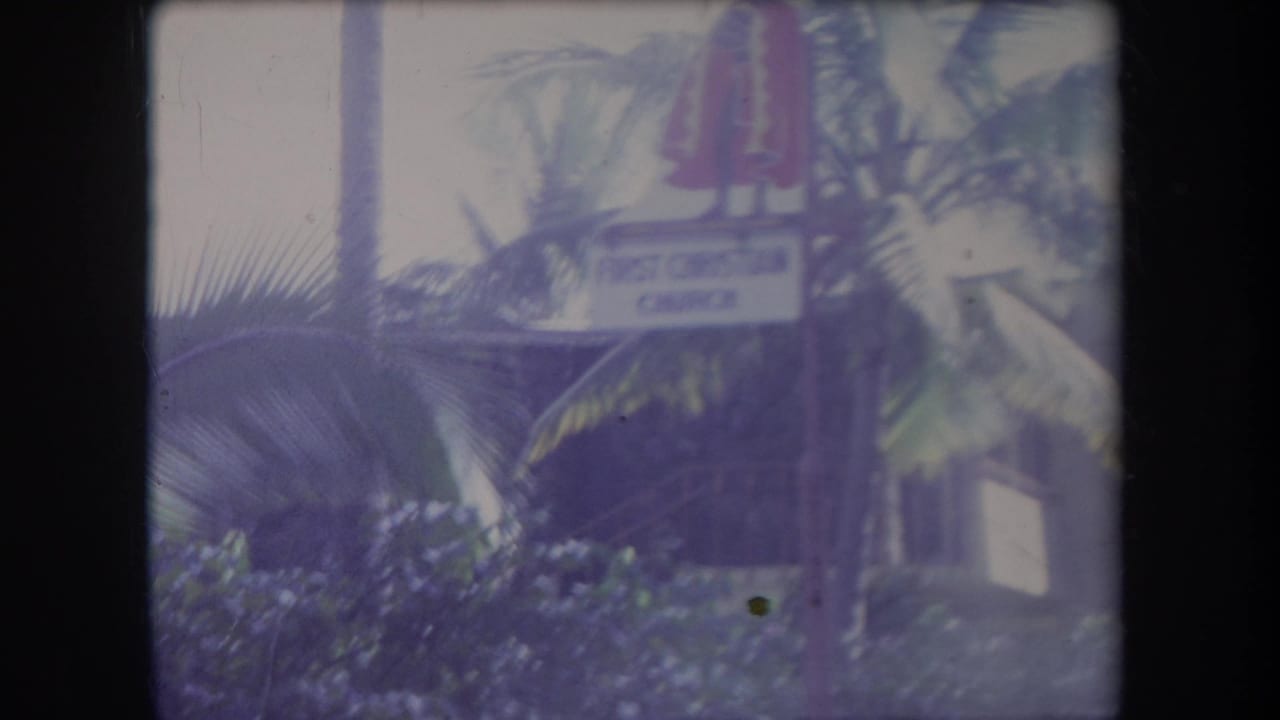 1969 Hawaii 8mm frame showing palm fronds, a stone building, a First Christian Church sign, and a red statue-like figure above the sign.
