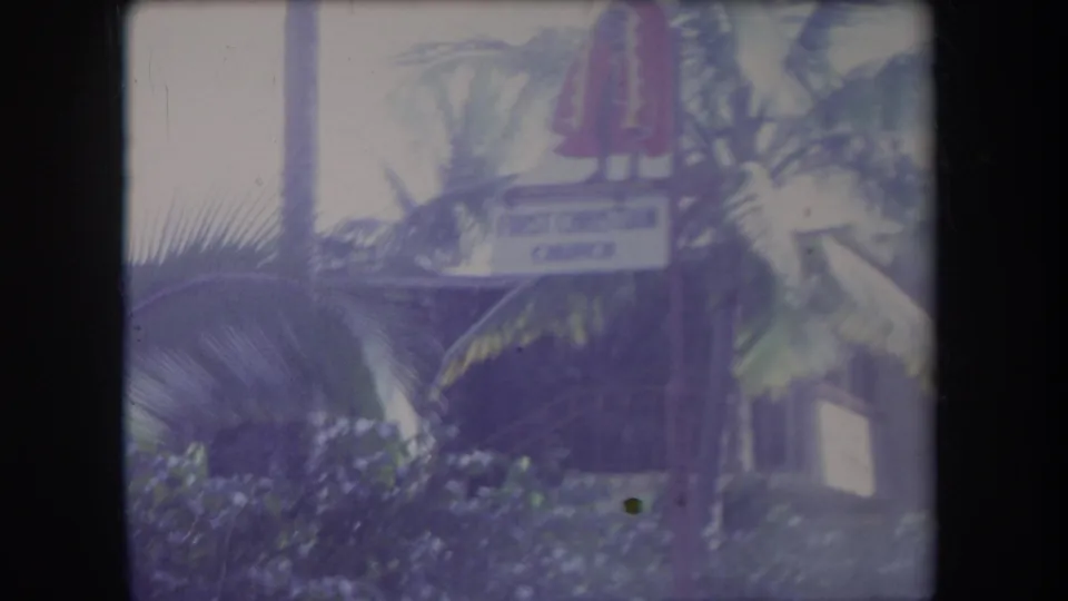 1969 Hawaii 8mm frame showing palm fronds, a stone building, a First Christian Church sign, and a red statue-like figure above the sign.