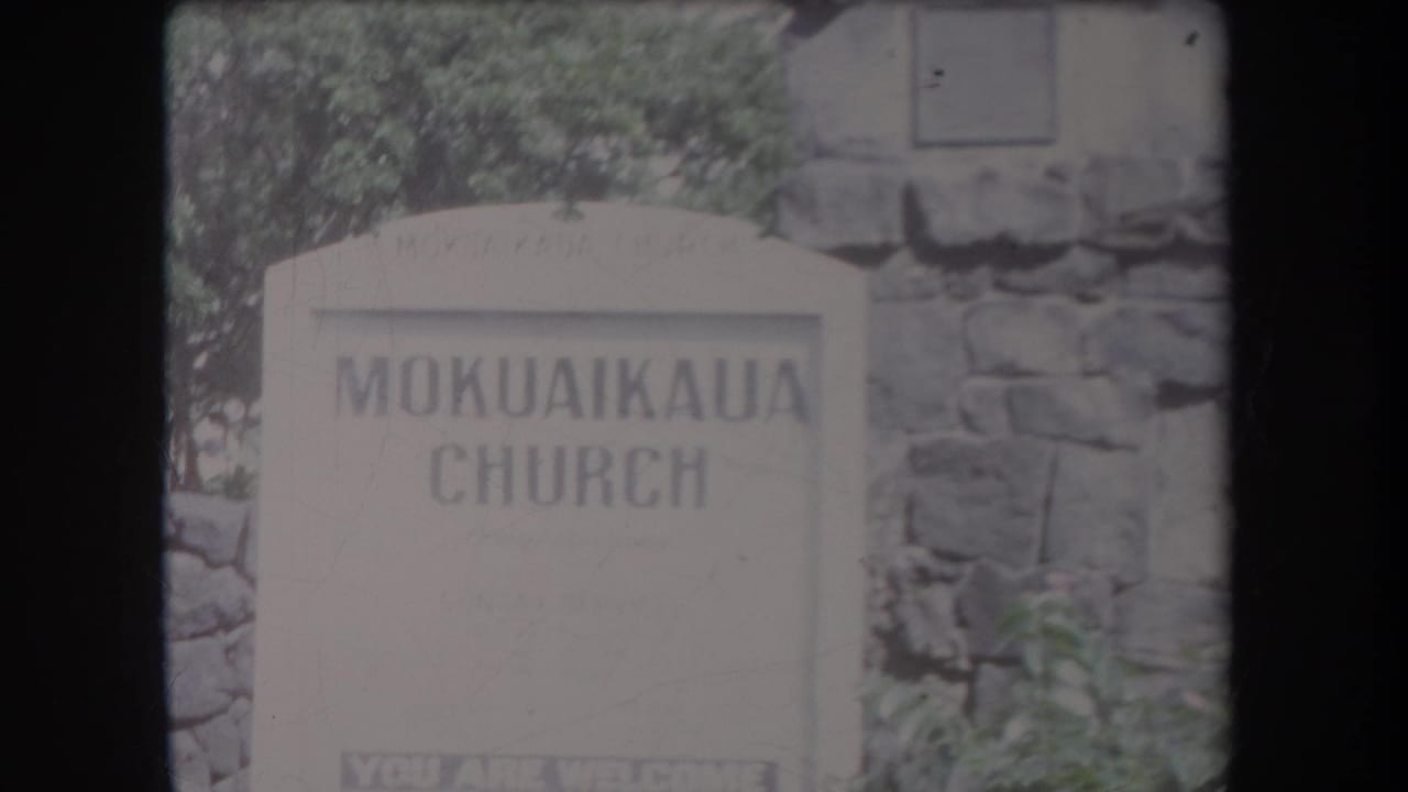 MokuaiKAUA Church sign in front of a stone wall and greenery.
