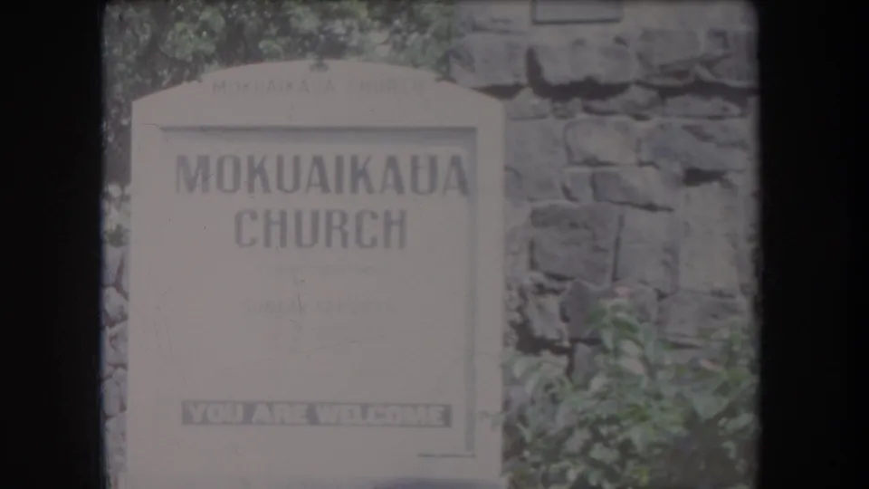 1969 Hawaii 8mm view of the MokuaiKAUA Church sign with rough masonry and leafy plants around it.