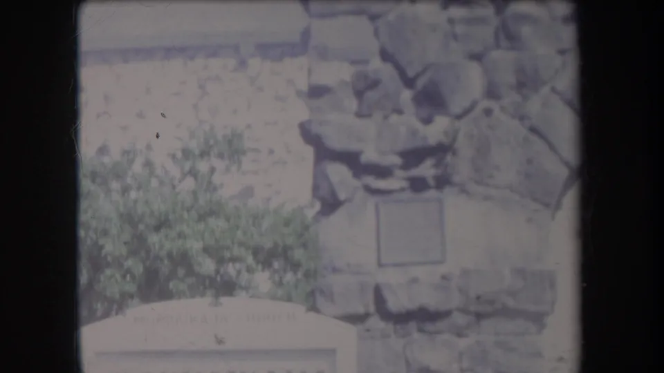 1969 Hawaii frame showing the lower edge of the MokuaiKAUA Church sign beneath stonework, foliage, and a small wall fixture.