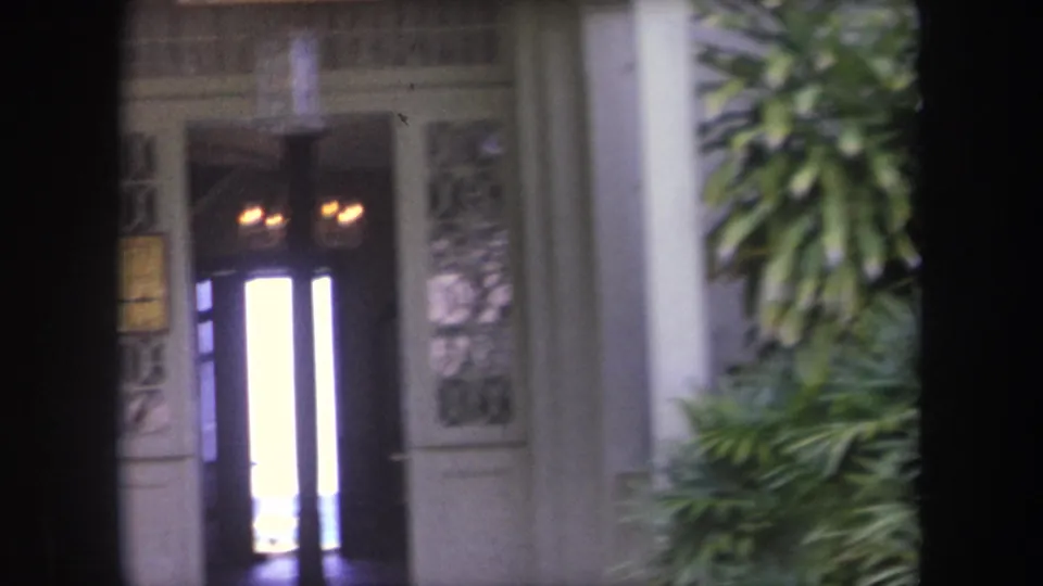 1969 Hawaii home-movie frame showing an ornate doorway, bright interior light, hanging fixture, and surrounding foliage.
