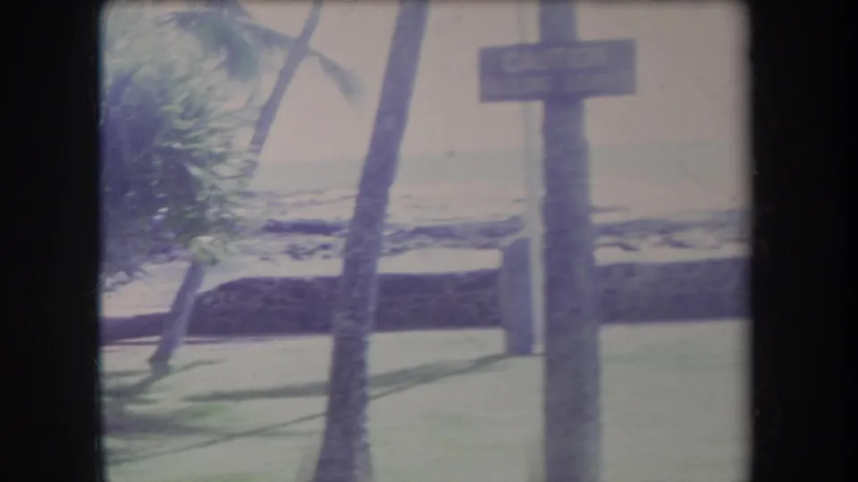 1969 Hawaii 8mm view of a wooden signpost near palms, lawn, and a hazy beach edge.