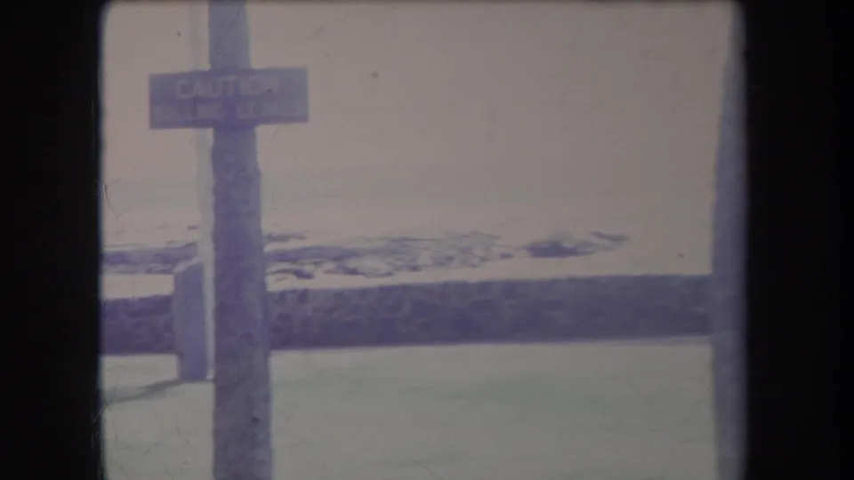 1969 Hawaii frame showing a wooden caution sign on a post, with lawn, beach, and pale ocean beyond.