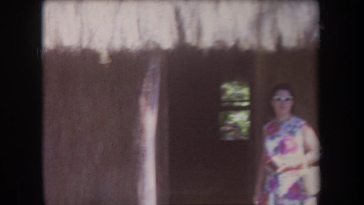 1969 Hawaii home-movie frame showing an adult woman in a floral dress and sunglasses near the shadowed entrance of a thatched structure.