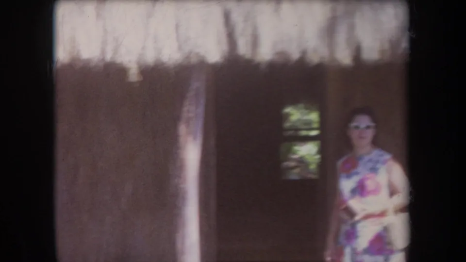 1969 Hawaii home-movie frame showing an adult woman in a floral dress and sunglasses near the shadowed entrance of a thatched structure.