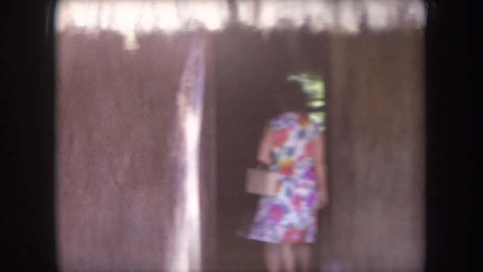 1969 Hawaii home-movie frame showing the woman from behind as she moves through the shadowed thatched structure with a handbag.