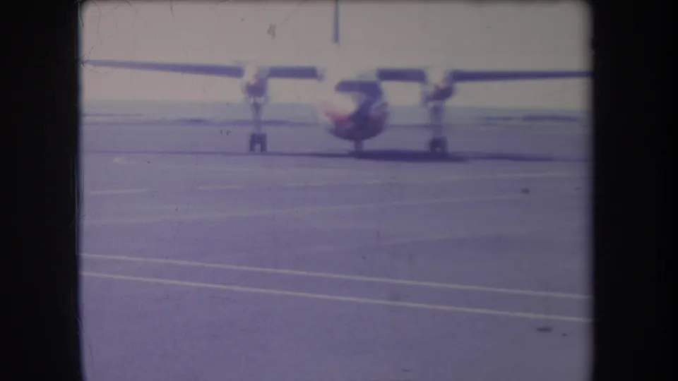 1969 Hawaii home-movie frame of a small twin-prop aircraft on a runway, with broad pavement and faint painted lines in front.