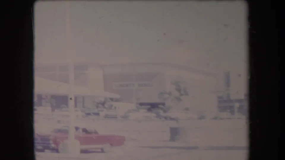 Wide faded 1969 Hawaii airport-area frame with low buildings, flags, parked cars, landscaping, and indistinct signage in the background.