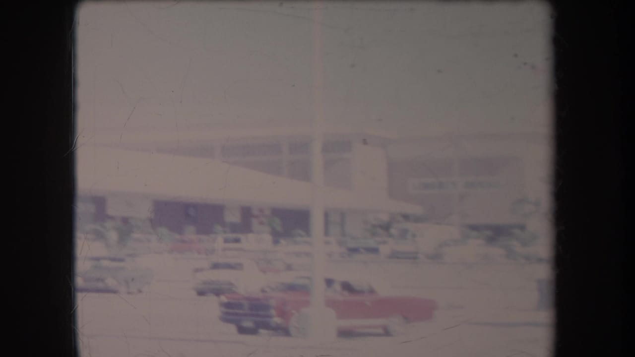 1969 Hawaii home-movie frame showing classic cars in a parking lot outside a low commercial building, with faded color and film scratches.