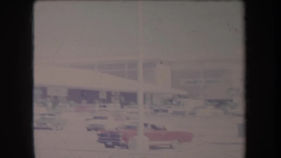 1969 Hawaii home-movie frame showing classic cars in a parking lot outside a low commercial building, with faded color and film scratches.