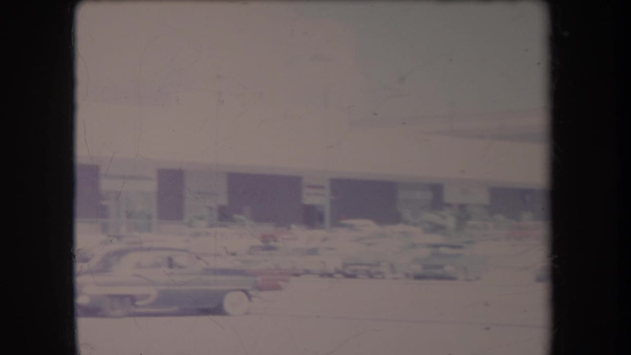 Parking lot scene with several cars in front of a low building and dark storefront bays.