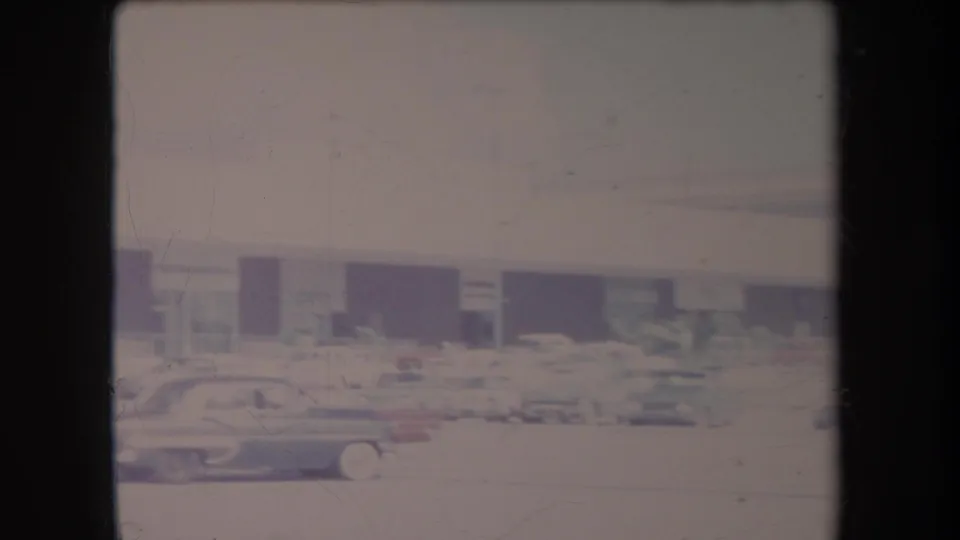 1969 Hawaii frame of parked cars and dark storefront openings across a lot, recorded in soft, faded 8mm color.