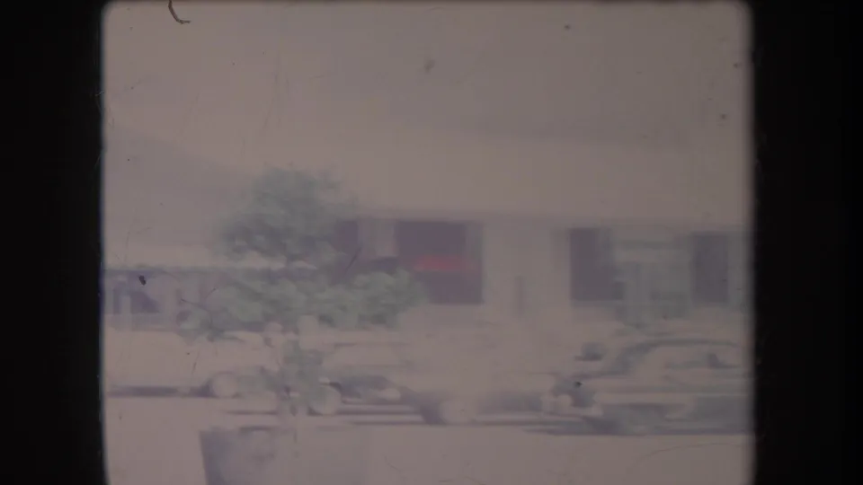 1969 Hawaii home-movie view of cars beside a low storefront, with a tree at left and heavy faded highlights.