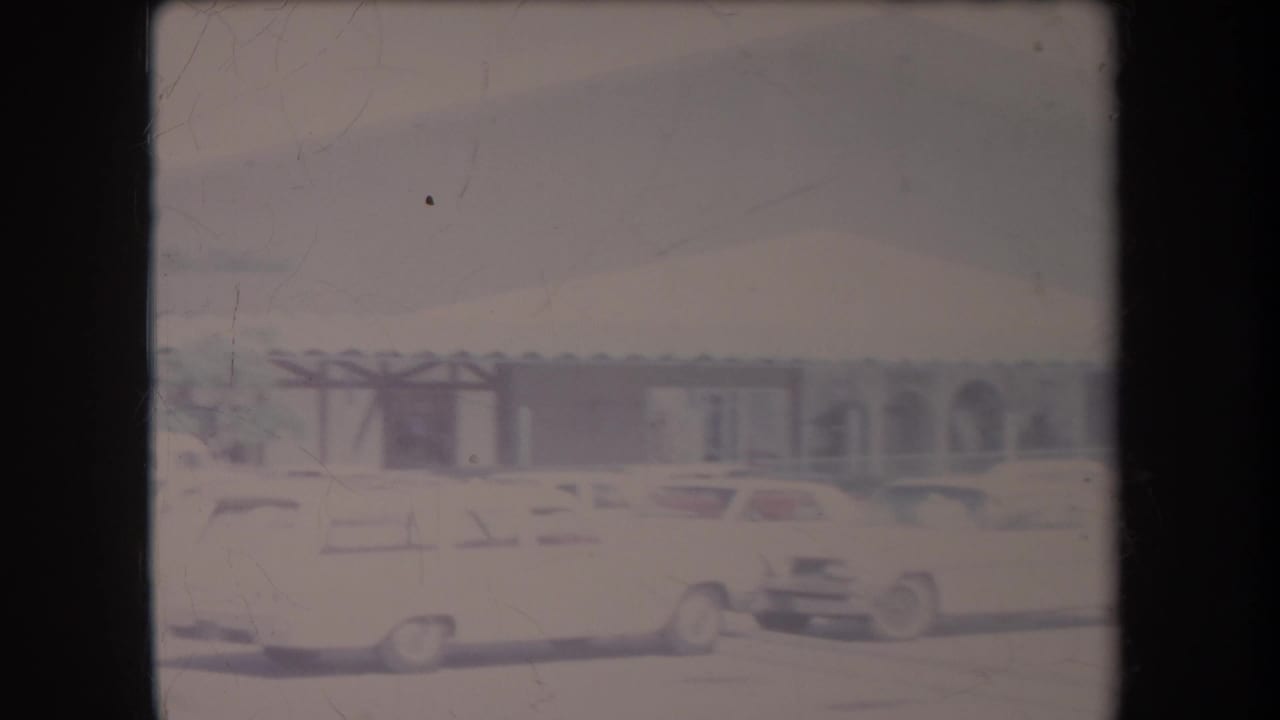 1969 Hawaii home-movie frame showing a bright parking lot, parked cars, and a low commercial building with palms nearby.