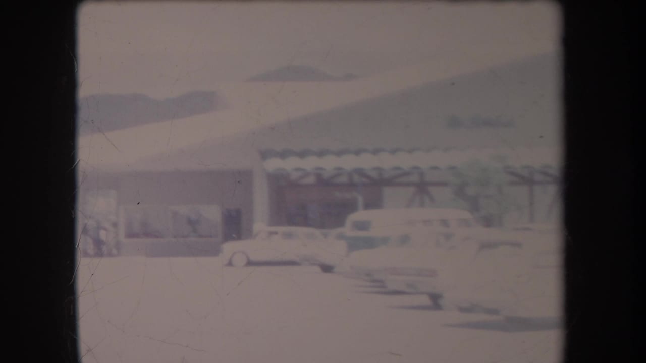 Cars parked near a low building with palm trees and a covered roofline.