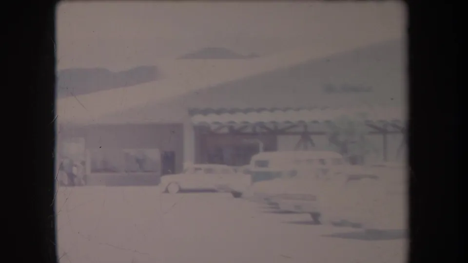 1969 Hawaii frame with late-1960s cars, palms, and a low commercial building seen across a bright parking lot.