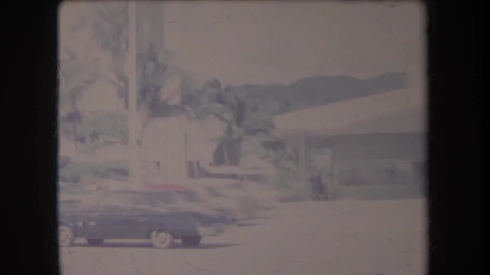 1969 Hawaii home-movie view of a dark car crossing the foreground near palms and a low modernist building.