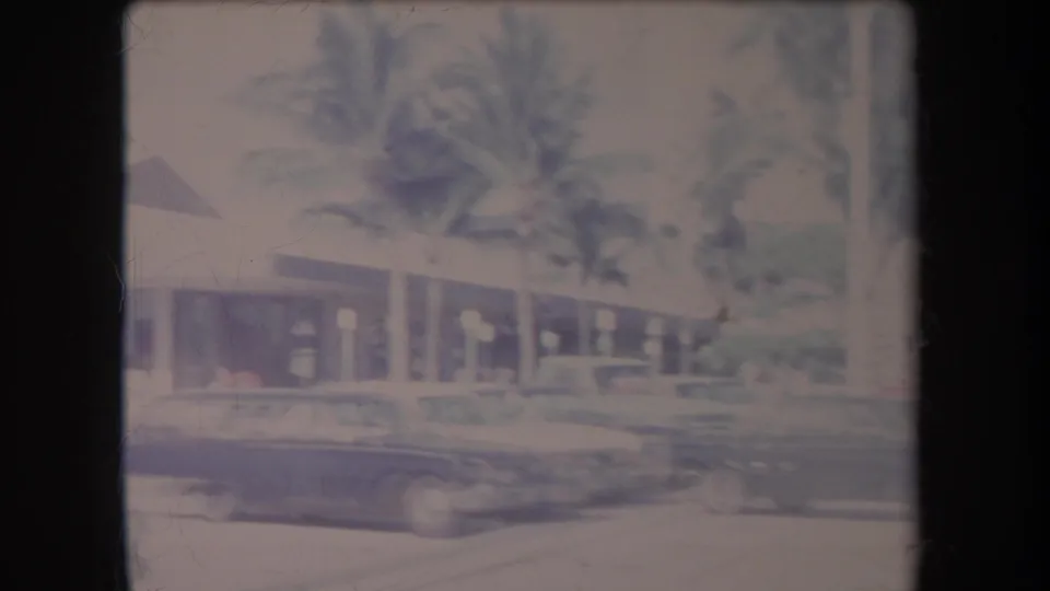 1969 Hawaii frame showing palms over a low commercial building, with cars moving or parked along the foreground lot.