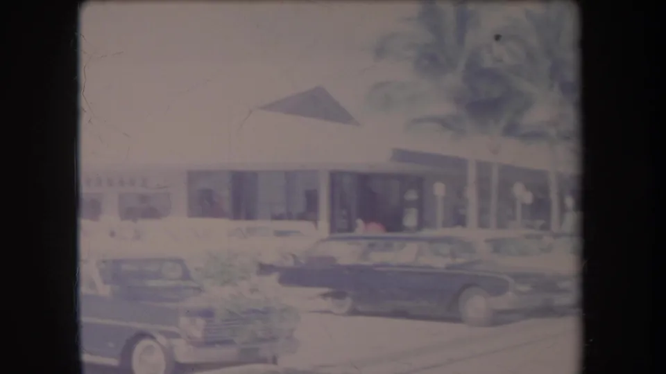 1969 Hawaii 8mm frame showing classic cars in a bright parking lot outside a low commercial building with a palm tree.