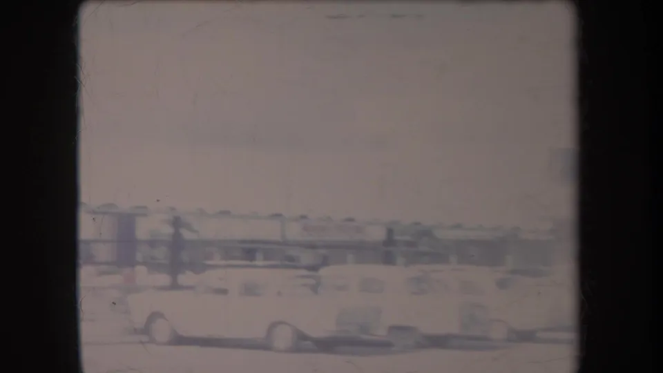 Faded 1969 Hawaii home-movie frame with a light-colored classic car, railings, open pavement, and a low roadside structure.
